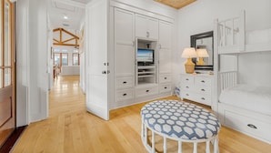 Une pièce avec un plafond en bois, des murs blancs, un lit superposé, une table ronde recouverte d’une nappe à motifs et des placards blancs intégrés, dont l’un abrite un téléviseur.