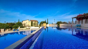 Piscina azul cristalina com borda azulejada, cercada por um edifício branco com varanda e uma pérgula com flores vermelhas.