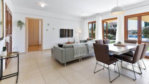 A modern living room with a grey sofa, a dining table with chairs, a flat-screen TV, and large windows offering a view of the outdoors.