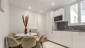 A modern kitchen with white cabinets, a black countertop, and a built-in microwave. There is a round dining table with four chairs and a vase with dried plants. - Sweett | Wagram Deluxe XI (Paris)