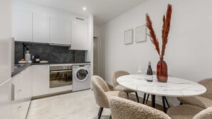 A modern kitchen with white cabinets, a built-in oven, and a washing machine. There is a round dining table with chairs and a vase with pampas grass. - Sweett | Wagram Deluxe XI (Paris)