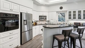 A modern kitchen with white cabinetry, stainless steel appliances, a central island with bar stools, and a window providing natural light. - Single Level Vacation Home w/ Private Pool (Hilton Head Island)