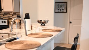 Une cuisine avec un plan de travail en marbre, trois sets de table en fibres tressées, un robinet noir, une machine à café et un tableau encadré accroché au mur.