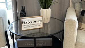 Une table à plateau en verre, agrémentée d’un panneau d’accueil, d’une plante en pot et d’un livre intitulé « La vie n’a pas besoin d’être parfaite pour être merveilleuse », dans un salon moderne avec un canapé beige et une table basse en verre.