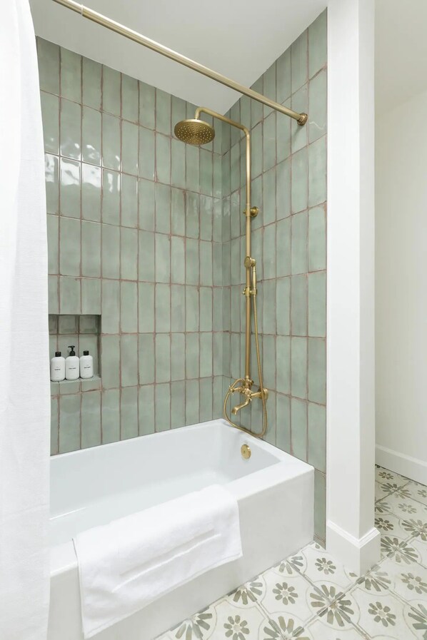 Un baño con una bañera blanca, una ducha con cabezal dorado y paredes cubiertas de azulejos verdes.