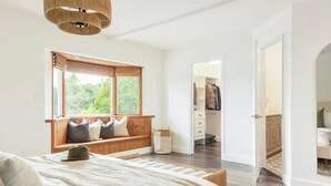 Ein Schlafzimmer mit einem großen Bett, einer hölzernen Bank mit Kissen, einem Fenster mit Blick ins Grüne, einem begehbaren Kleiderschrank und einer Tür zu einem weiteren Raum.