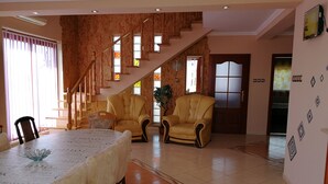 A spacious living room with a high ceiling, a staircase leading to an upper floor, a dining area with a table and chairs, and a large window with curtains.