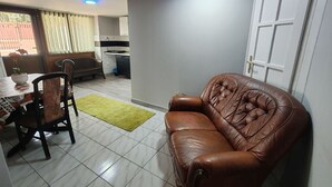 A living room with a brown leather sofa, a dining table with chairs, and a kitchen in the background.