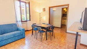 Un salon avec un canapé bleu, une table à manger entourée de six chaises, une cuisine visible à travers une porte ouverte et un téléviseur posé sur un meuble.