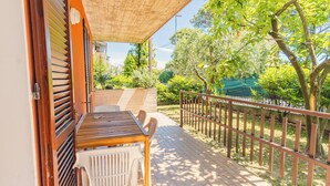 Une terrasse extérieure avec une table et des chaises en bois, une rambarde en métal et une vue sur un jardin avec des arbres et une clôture.
