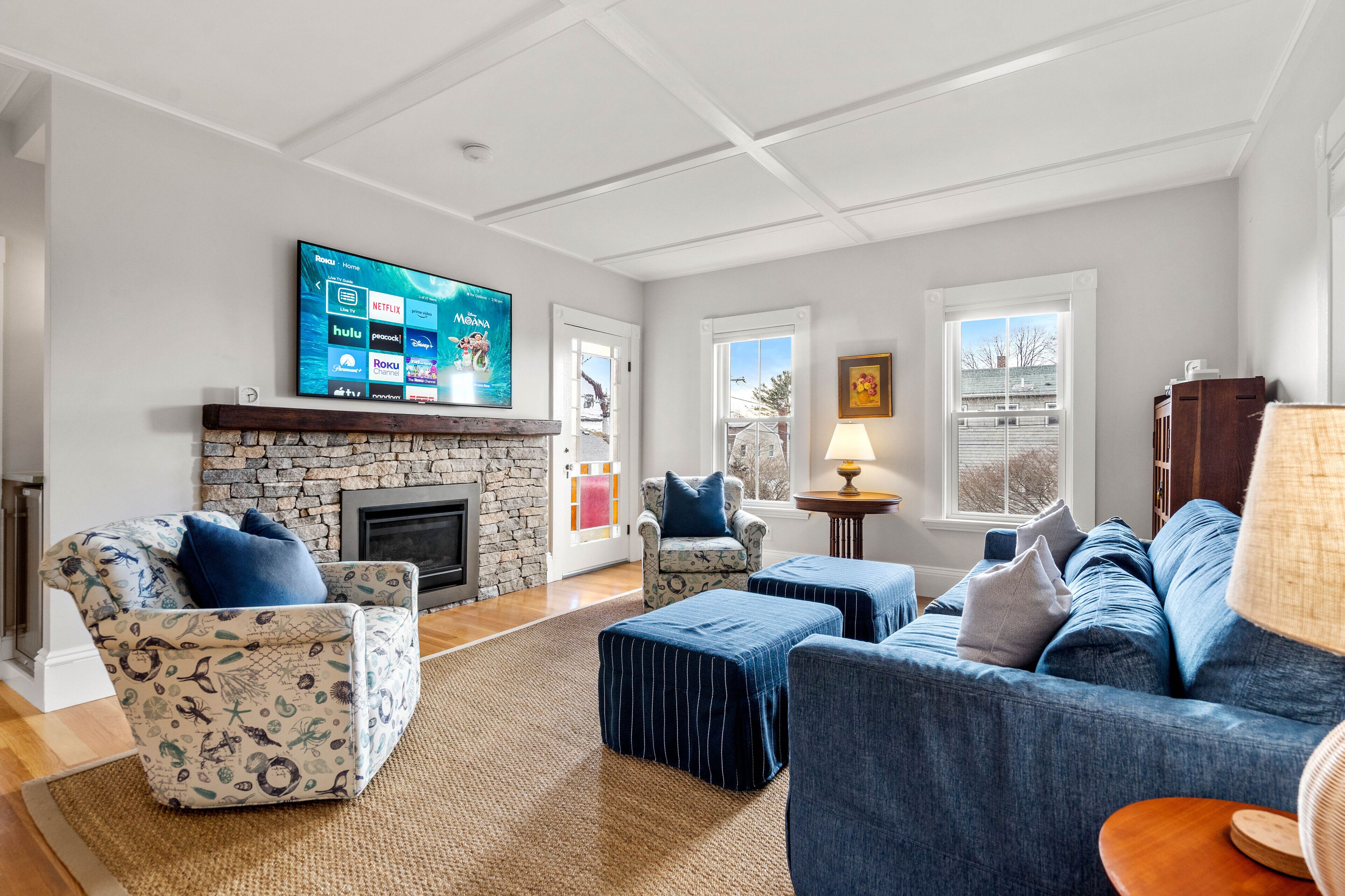 Ein Wohnzimmer mit Kamin, einem flachbildfernseher, der darüber angebracht ist, und einer Steinwand. Es gibt zwei Sofas, einen gemusterten Sessel und einen kleinen Tisch mit einer Lampe.