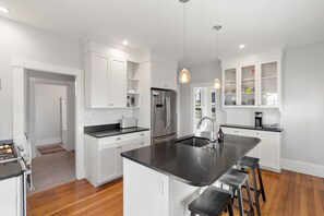 A modern kitchen with white cabinetry, a central island with bar stools, stainless steel appliances, and pendant lighting. - The Mariner · Walk to Beach+Rocky Neck · Fenced Backyard (Gloucester)