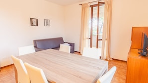 Une pièce avec une table en bois, des chaises blanches, un canapé gris et un téléviseur posé sur un meuble en bois.