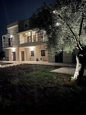 Um edifício de dois andares com varandas, uma árvore em primeiro plano e iluminação externa.