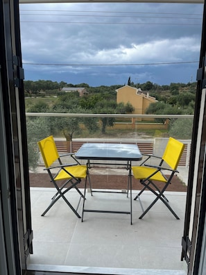 Varanda com mesa e duas cadeiras amarelas, vista para uma área residencial com casas e vegetação.