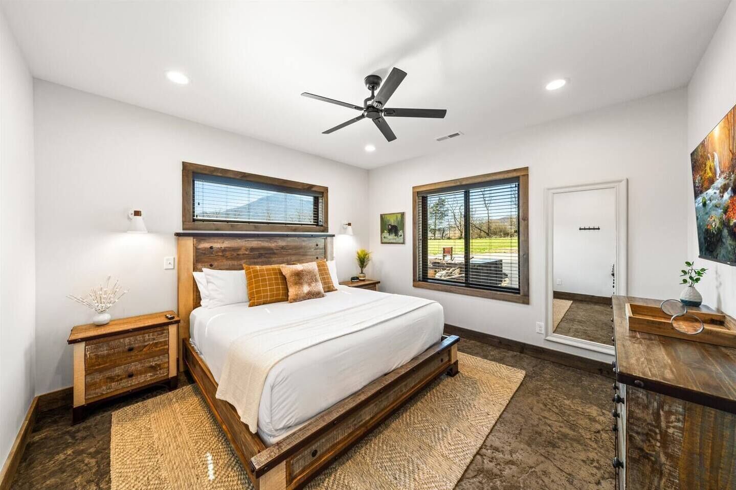 Une chambre avec un grand lit, une tête de lit en bois, deux tables de chevet, un ventilateur de plafond et une vue sur l’extérieur grâce à deux fenêtres.