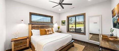 Une chambre avec un grand lit, une tête de lit en bois, deux tables de chevet, un ventilateur de plafond et une vue sur l’extérieur grâce à deux fenêtres.