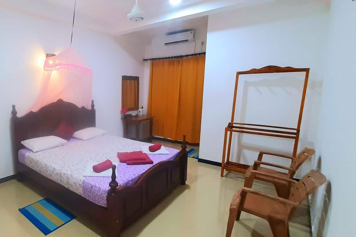 A bedroom with a bed, a wooden wardrobe, a desk with a chair, and a standing fan.