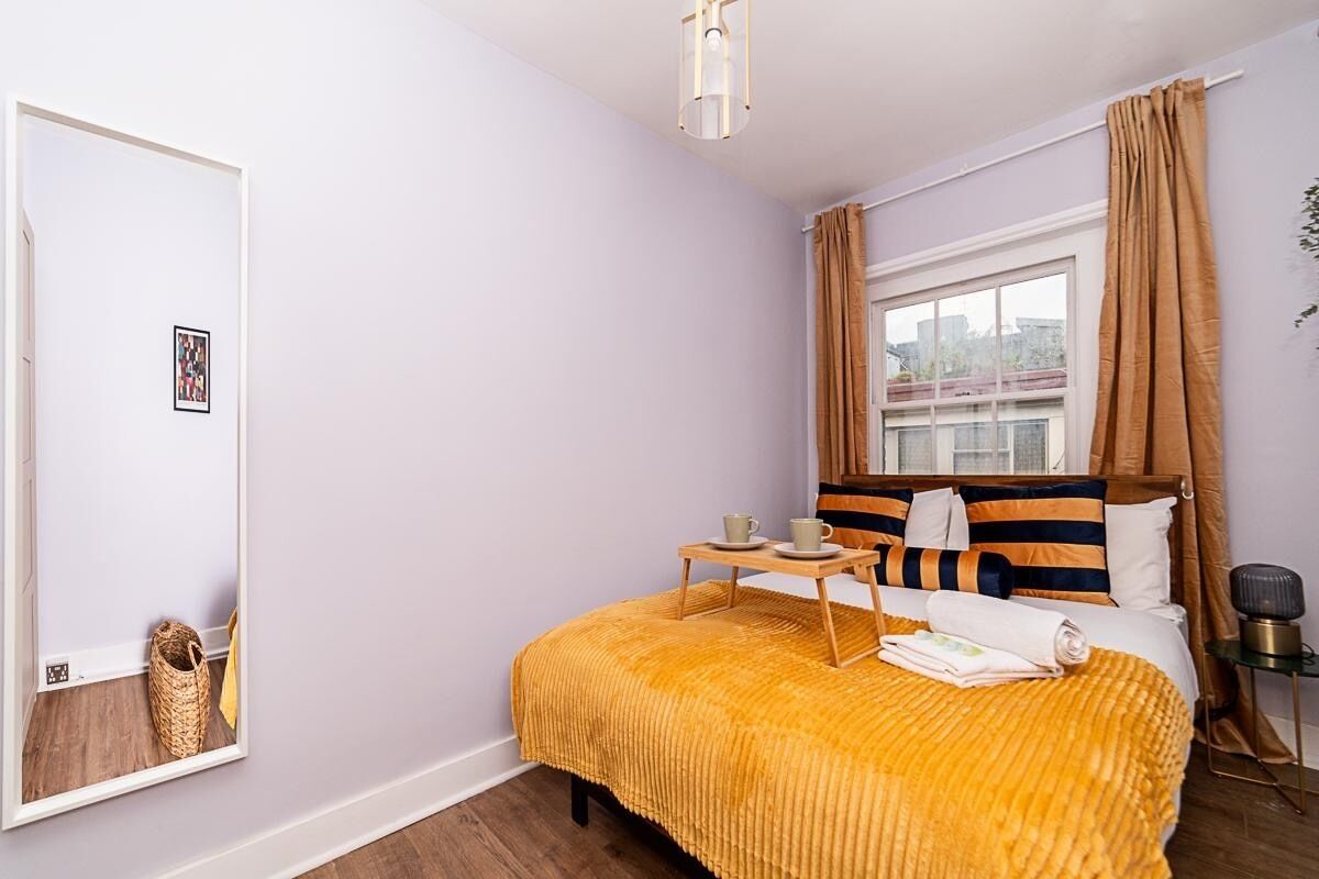 Quarto com cama, mesa de cabeceira, janela com cortinas, espelho e luminária na parede.
