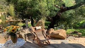 Une terrasse avec un fauteuil de relaxation en bois, une table ornée d’un vase de tournesols et une petite table décorative blanche.