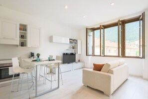 Une cuisine moderne avec des meubles blancs, un coin repas avec une table et des chaises blanches, et un canapé offrant une vue sur la ville grâce à de grandes fenêtres.