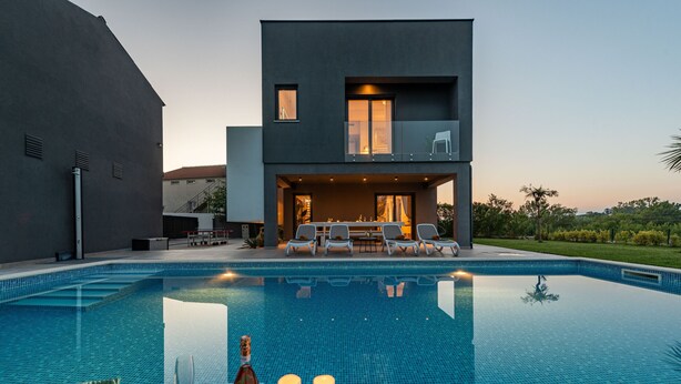 Una casa moderna con piscina, mobiliario exterior e iluminado balcón.