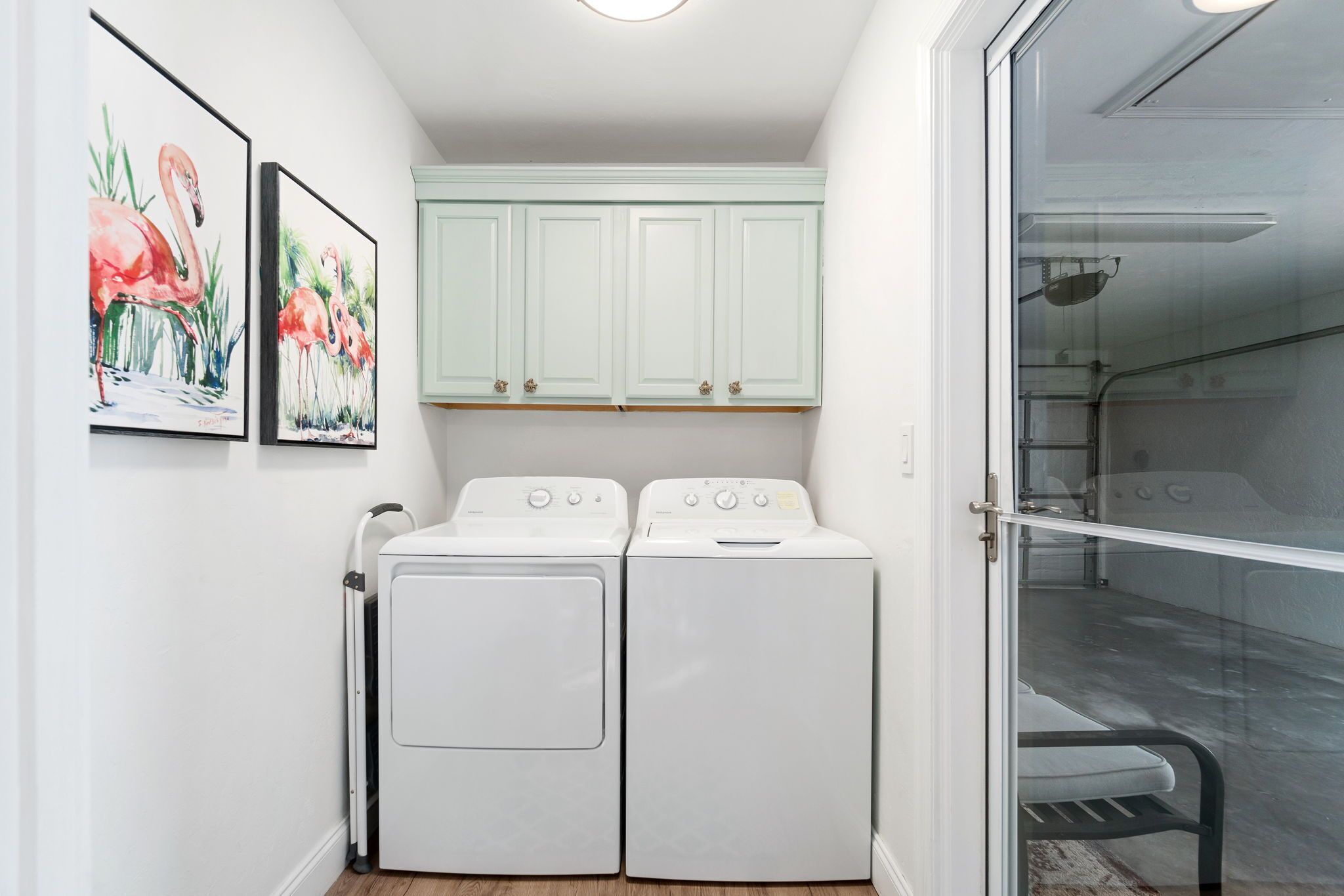 A laundry room with two white washing machines, a small cabinet above them, and two framed paintings on the wall.
