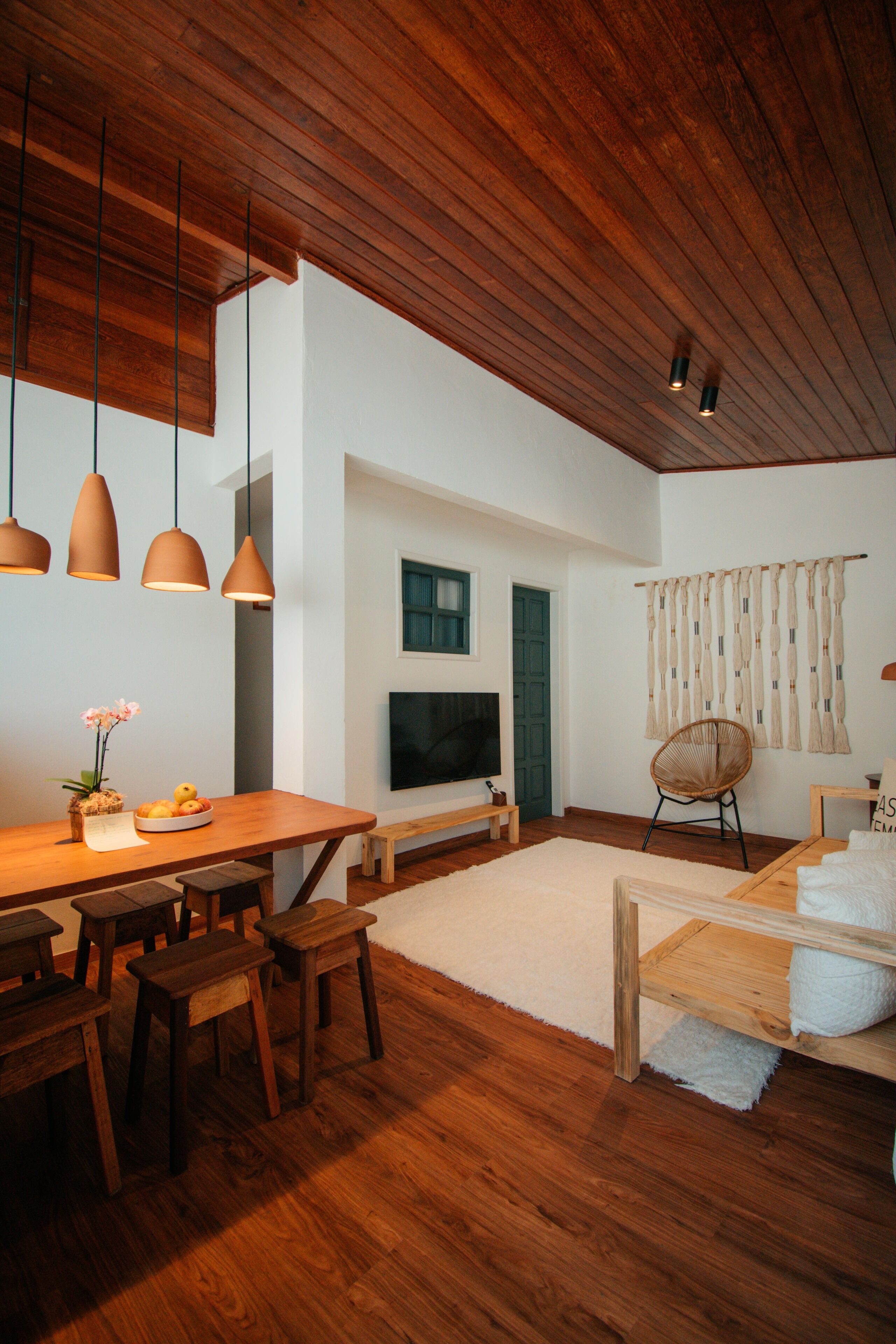 Uma sala de estar moderna com piso de madeira, área de jantar com mesa e bancos de madeira, televisão fixada na parede e uma janela grande com cortinas.