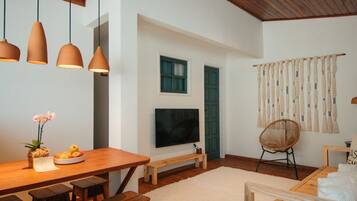 Uma sala de estar moderna com piso de madeira, área de jantar com mesa e bancos de madeira, televisão fixada na parede e uma janela grande com cortinas.