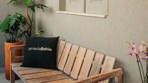 A wooden bench with a black pillow, a potted plant, and a window with shutters. - Pousada Girassol Morro (Cairu)