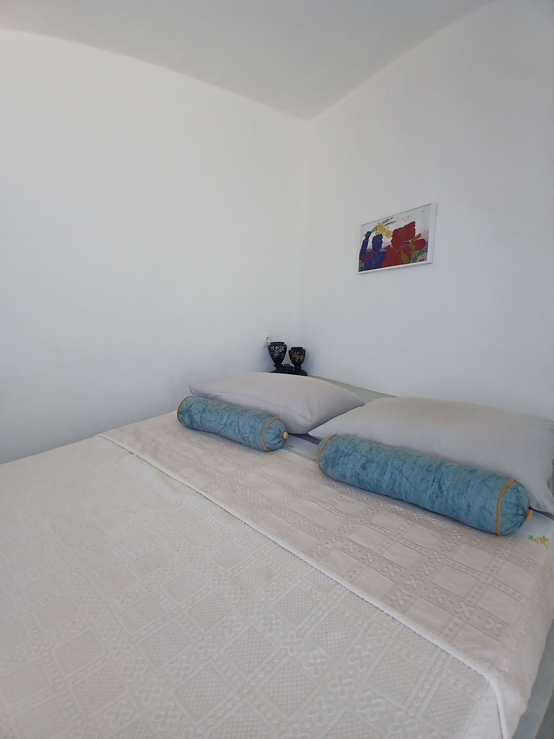 Ein Einzelbett mit gemustertem Bettbezug und zwei Kissen, ein kleiner Tisch mit zwei Dekorationsobjekten sowie ein farbenfrohes Bild an der Wand.