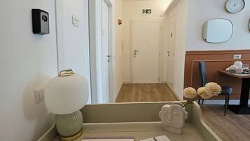 A hotel room with a desk, a lamp, a vase with flowers, a clock, and a door with an emergency exit sign.