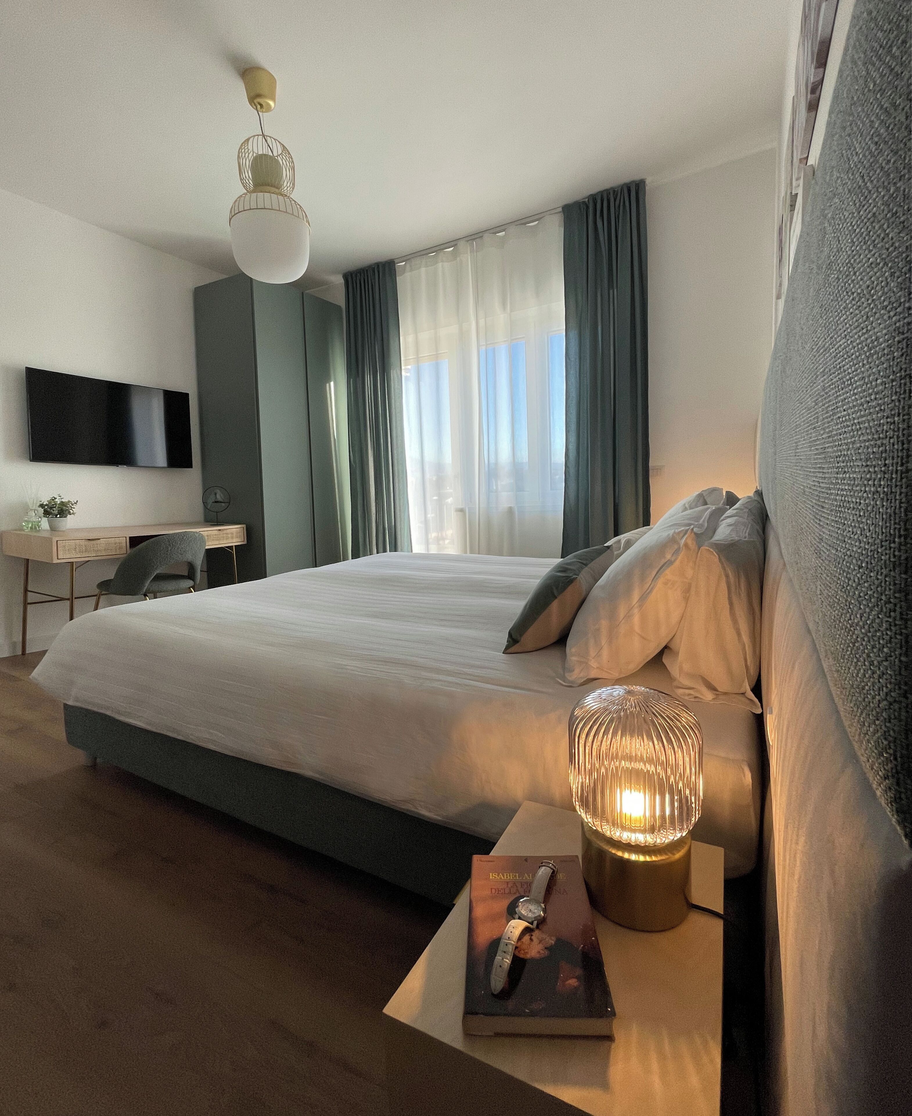 A modern hotel room with a large bed, a desk with a chair, a flat-screen TV, and a bedside table with a lamp and book.