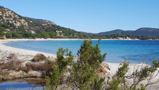 A coastal landscape with a sandy beach, clear blue water, and a small pond surrounded by greenery.