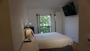 Ein Schlafzimmer mit einem Bett, einem Schreibtisch mit Lampe, einem an der Wand montierten Fernseher und einem Fenster mit Blick auf Bäume.