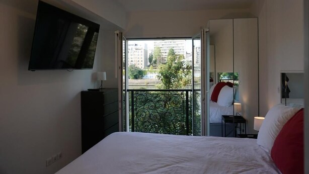 Ein Hotelzimmer mit Bett, Wandfernseher, Balkon mit Blick auf Gebäude und Bäume sowie einem Kleiderschrank.