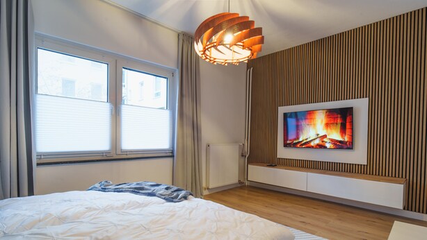 Un dormitorio con una cama, un televisor montado en la pared y un ventanal con cortinas.
