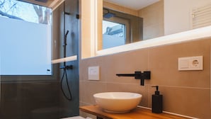 Un baño moderno con ducha envuelta en vidrio, un inodoro blanco, un tocador de madera con un lavamanos blanco y un espejo sobre el tocador.
