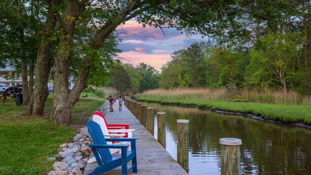 A wooden pier with Adirondack chairs, a calm river, and trees in the background.
