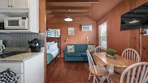 A compact kitchen with a microwave, a sink, and a countertop with utensils. There is a dining area with a wooden table and chairs, and a bunk bed with a green sofa.