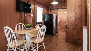 A wooden cabin interior with a dining table, four white chairs, a black refrigerator, a television mounted on the wall, and a window with blinds.