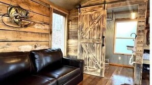 A rustic interior with a black leather sofa, a cowhide rug, wooden walls, and a large wooden door. - Fishing Cabin Getaway (Grant)