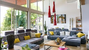 Ein geräumiges Wohnzimmer mit großen Fenstern, grauen Sofas mit gelb-weißen Kissen, einem hölzernen Couchtisch und roten Pendelleuchten.