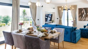 A modern living room with a dining area, featuring a blue sofa, wooden dining table, and pendant lights. - Villa Lux Zadarvillas (Zemunik Donji)