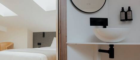 A modern bathroom with a skylight, a large oval mirror, a white sink, and two bottles of shampoo.