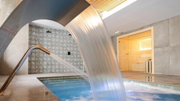 Una piscina interior moderna con una cascada, una zona de sauna y un muro con azulejos.