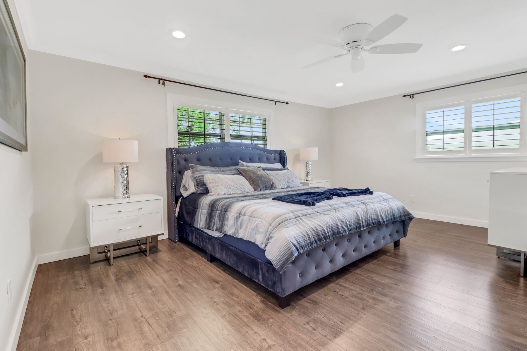 A bedroom with a large bed, two bedside tables, a ceiling fan, and windows with blinds.