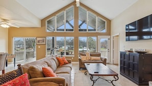 A spacious living room with a high ceiling, large windows, a sofa set, a coffee table, and a TV mounted on the wall. - Stunning Lake Home Get Away on The Water! (Gun Barrel City)