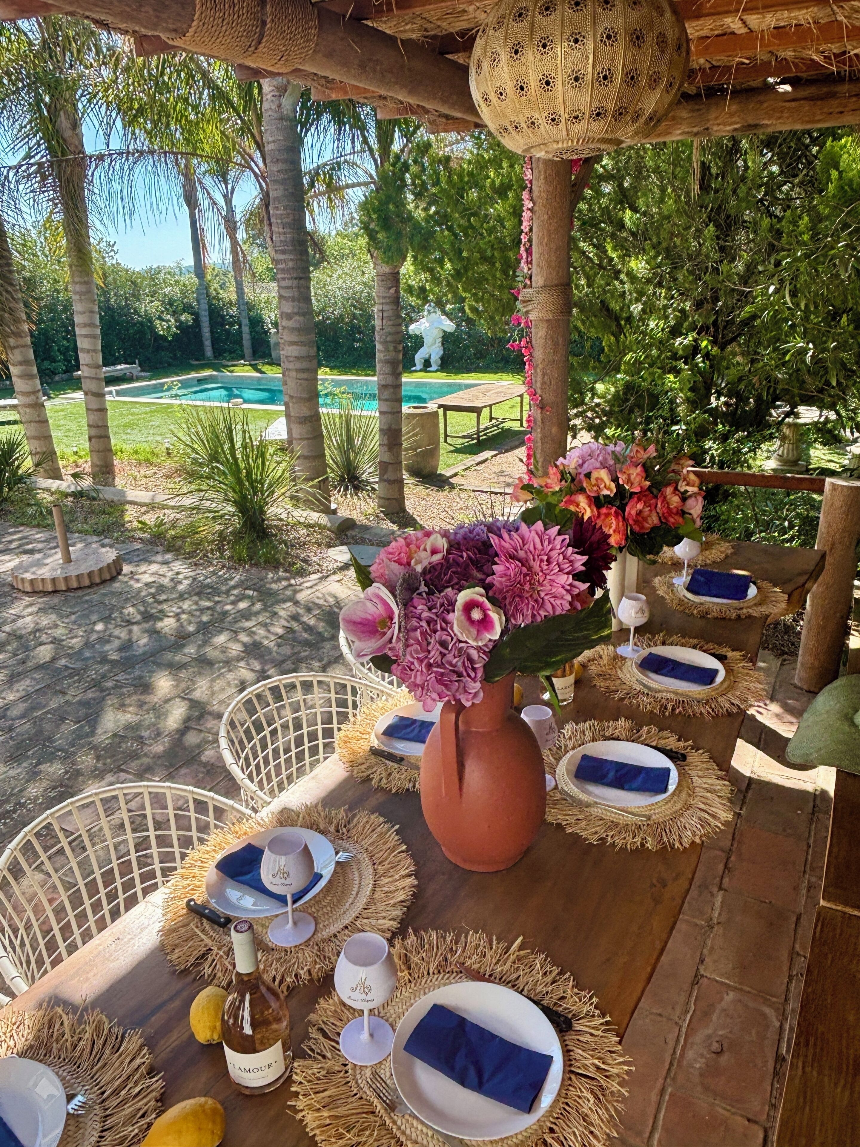 An outdoor dining area with wicker furniture, a large vase of flowers, and a view of a pool and greenery.