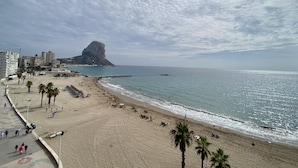 Una playa con un promontorio rocoso, palmeras y un paseo marítimo con gente caminando.